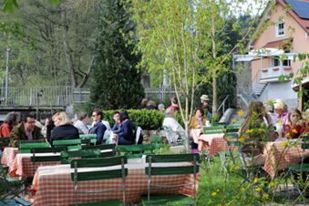People sitting at tables with orange and white checkered tablecloths in the garden area of Landgasthof Hotel Hammerm&uuml;hle surrounded by trees and flowers