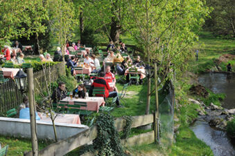 Outdoor dining area at Landgasthof Hotel Hammerm&uuml;hle with green chairs, checkered tables, and people seated near a small river