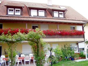 Two-story building with red flower boxes on the balcony, white chairs and tables outside, and green plants at G&auml;stehaus Rita