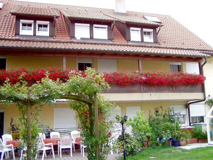 Two-story building with red flower boxes on the balcony, white chairs and tables outside, and green plants at G&auml;stehaus Rita