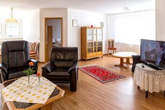Bright lounge at G&auml;stehaus Rita with leather armchairs, wooden furniture, a TV, round and hexagonal tables, and a red rug