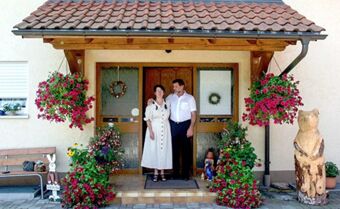 A man and woman standing at the entrance of G&auml;stehaus Rita with hanging flower baskets and wooden bear carving nearby