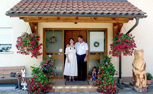 A man and woman standing at the entrance of G&auml;stehaus Rita with hanging flower baskets and wooden bear carving nearby