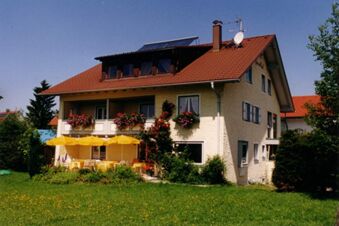 Two-story yellow building with red roof, flower boxes on balcony, yellow umbrellas and chairs in garden of Pension G&auml;stehaus Alpenblick