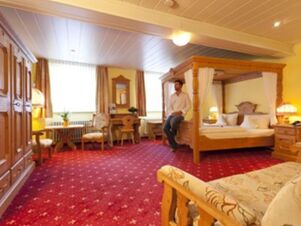 Room interior at Akzent Hotel Goldner Stern featuring a four-poster bed, wooden furniture, red patterned carpet, and a seated man