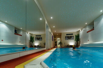 Indoor swimming pool area with blue water, large wall mirror, plants, benches, and artwork at Akzent Hotel Goldner Stern