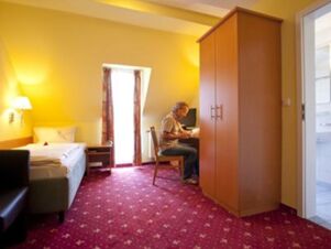 A man sitting at a desk in a yellow-walled room with a red patterned carpet and wardrobe at Akzent Hotel Goldner Stern