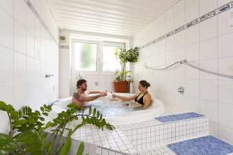 A man and woman in swimwear sitting in a white tiled hot tub clinking glasses inside Akzent Hotel Goldner Stern bathroom