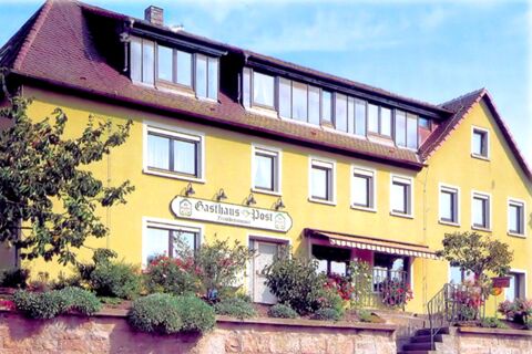 Die gelbe Fassade des Gasthauses Zur Post mit vielen Fenstern, Blumenbepflanzung und einem schr&auml;gen Dach an einem sonnigen Tag.