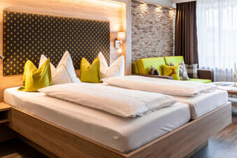 Modernes Hotelzimmer im Gasthof-Metzgerei St&ouml;berl mit zwei Betten, gr&uuml;nen Kissen und Sofa vor einer Steinwand und Fenster mit Gardinen.