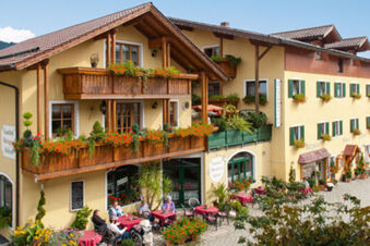 Ansicht der Fassade mit Balkon und blumengeschm&uuml;ckter Terrasse des Gasthof-Metzgerei St&ouml;berl mit Sitzgruppen und G&auml;sten im Au&szlig;enbereich.