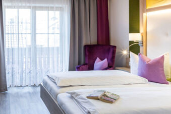 Helles und gem&uuml;tliches Zimmer mit Doppelbett, lila Sessel und Vorh&auml;ngen im Gasthof-Metzgerei St&ouml;berl mit Balkonfenster.