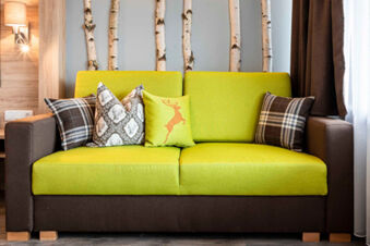 Gem&uuml;tliche gr&uuml;ne Couch mit dekorativen Kissen vor einer Wand mit Birkenst&auml;mmen im Zimmer des Gasthof-Metzgerei St&ouml;berl.