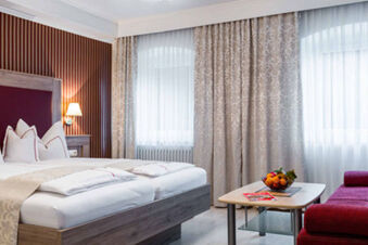 Helles und gem&uuml;tliches Hotelzimmer mit Doppelbett, rotem Sofa und Fenster mit Gardinen im Gasthof-Metzgerei St&ouml;berl.