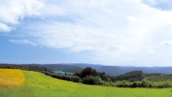 Rolling green and yellow fields with distant forested hills under a partly cloudy sky near Hotel Post Nordhalben