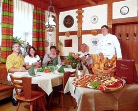 Four guests seated at a table with green napkins and flowers, and a chef standing beside a table of bread and appetizers at Hotel Post Nordhalben