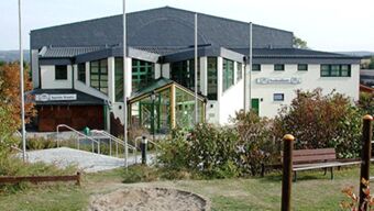 White multi-story building with dark roof and green windows labeled Hotel Post Nordhalben surrounded by bushes and seating area