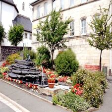 A landscaped garden area with flowers, bushes, trees, stone water feature, and a red bench at Hotel Post Nordhalben