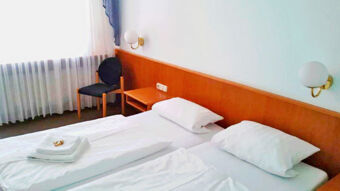 Schlicht eingerichtetes Hotelzimmer im Gasthof Waldbrunner Hof mit zwei Einzelbetten, einem Stuhl und einem Fenster mit Gardinen f&uuml;r Tageslicht.