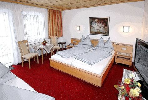 Double bed with blue bedding, wooden furniture, red carpet, table with two chairs, and a TV in Pension Haus Pilch room