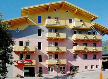 Multi-story building Pension Haus Pilch with beige balconies decorated with flowers and a yellow wooden roof under a blue sky