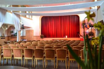 Rows of empty wooden chairs face a stage with a closed red curtain in a well-lit room at Posthotel Johannesberg