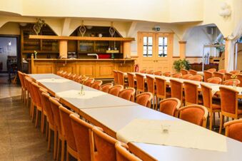 Dining area with long tables and orange chairs near a wooden bar inside Posthotel Johannesberg