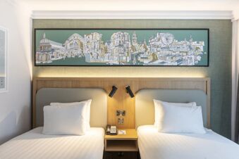 Two single beds with white linens and pillows in a room at Holiday Inn London-Regent's Park with cityscape artwork above