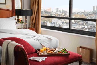 Bright hotel room at Millennium Hotel London Knightsbridge with a white bed, red chaise lounge, breakfast tray, and city view through large window