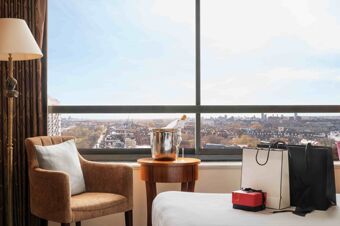 Room interior at Millennium Hotel London Knightsbridge with armchair, standing lamp, side table with ice bucket and champagne bottle, and shopping bags by window showing cityscape outside