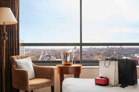 Room interior at Millennium Hotel London Knightsbridge with armchair, standing lamp, side table with ice bucket and champagne bottle, and shopping bags by window showing cityscape outside