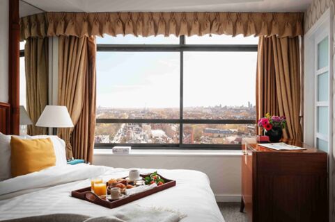 A hotel room at Millennium Hotel London Knightsbridge with a bed, breakfast tray, large window, brown curtains, and a wooden cabinet with flowers