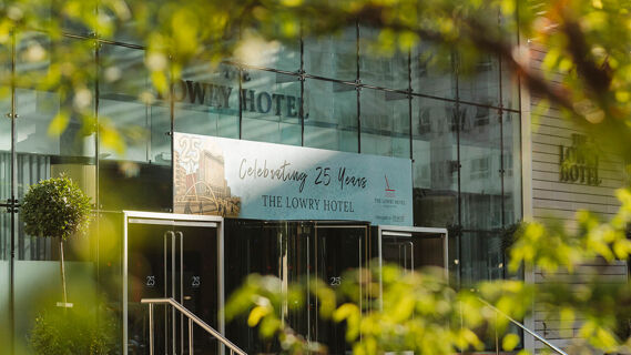 The glass entrance of The Lowry Hotel Manchester with a banner celebrating 25 years and green foliage in the foreground