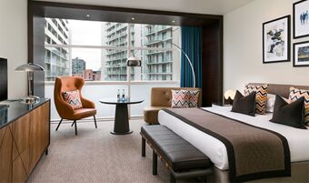 Hotel room at The Lowry Hotel Manchester with a king bed, brown armchair, round table, and large city-view window