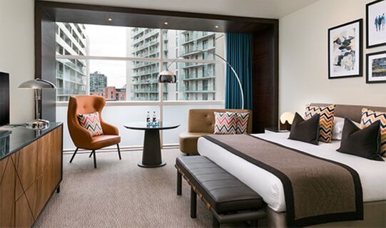 Hotel room at The Lowry Hotel Manchester with a king bed, brown armchair, round table, and large city-view window