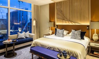 Modern hotel room at The Lowry Hotel Manchester with a large bed, blue bench, sofa, lamps, and city view at dusk