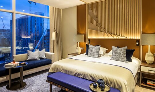 Modern hotel room at The Lowry Hotel Manchester with a large bed, blue bench, sofa, lamps, and city view at dusk