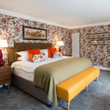 A double bed with white linens, orange pillows, floral cushions, green throw, and a yellow bench in a floral wallpapered room at Hotel du Vin Glasgow