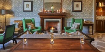 A sitting area with green armchairs, a lit fireplace, floral cushions, a wooden table with candles, and decorative wallpaper at Hotel du Vin Glasgow