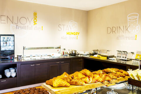 Croissants and assorted pastries on a wooden board in a breakfast area at B&B Hotel Bari Rond&ograve; with wall signs and buffet counters