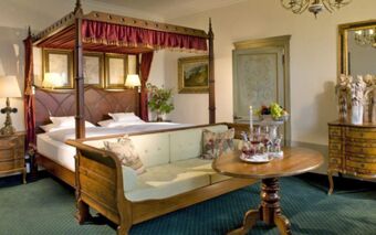 Four-poster bed with burgundy canopy, wooden sofa with cushions, round table with wine glasses, and vintage decor in Burghotel Auf Sch&ouml;nburg room