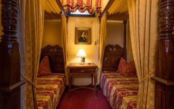 Two wooden carved canopy beds with red and gold patterned bedspreads and curtains flank a nightstand with a lamp at Burghotel Auf Sch&ouml;nburg