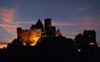 Burghotel Auf Sch&ouml;nburg illuminated at night on a hilltop with a dark sky and scattered pink clouds in the background