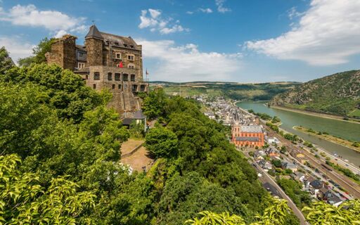 Burghotel Auf Sch&ouml;nburg castle on a hill surrounded by green trees overlooking a river, town, and railway tracks under a blue sky