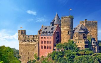 Burghotel Auf Sch&ouml;nburg castle with stone towers, a red brick building, greenery at the base, and two German flags flying above