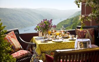 Outdoor breakfast table with drinks, flowers, and food set on scenic balcony at Burghotel Auf Sch&ouml;nburg overlooking river and hills