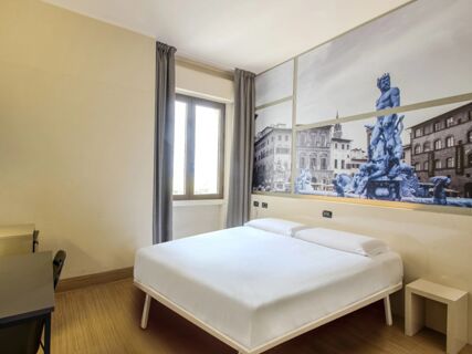 Bedroom with white bed, wooden floor, window with gray curtains, and wall mural of a fountain at B&B Hotel Firenze Novoli