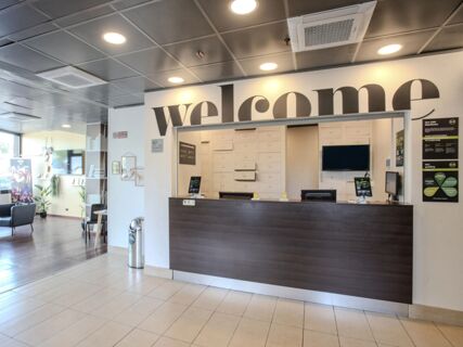 Reception desk with "welcome" sign above and informational posters visible in the lobby area of B&B Hotel Firenze Novoli