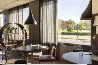A dining area at B&B Hotel Arezzo with round tables, chairs, pendant lamps, sheer curtains, and a circular plant stand near large windows