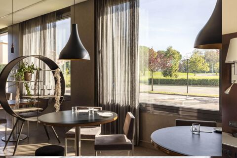 A dining area at B&B Hotel Arezzo with round tables, chairs, pendant lamps, sheer curtains, and a circular plant stand near large windows